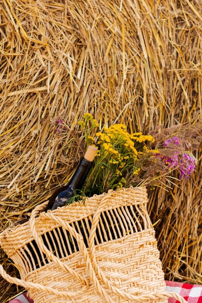 Bottle of red wine, two glasses and wildflowers in basket on the field and sheaf.