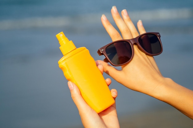Bottle of sunscreen lotion suncream packaging on the sandy\
beach sea sky background copy spacesunglasses on hand palmoil spa\
spf vacation on tropical paradise copyspacehello summer