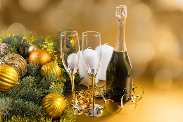 Bottiglia di champagne natalizio sigillato senza etichetta per celebrare la stagione in piedi con due eleganti flauti accanto a un ramo di pino decorato con palline d'oro con uno sfondo sfocato con copyspace