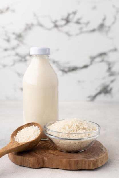 A bottle of vegetable rice milk and a bowl of rice on a wooden board with a spoonful of grains on a light background