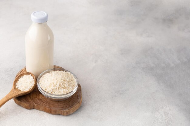 Una bottiglia di latte di riso vegetale e una ciotola di riso su una tavola di legno con un cucchiaio di cereali su uno sfondo chiaro