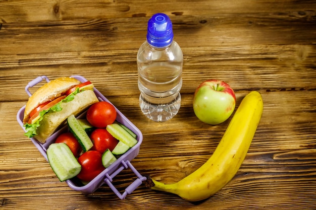 水のリンゴバナナのボトルと木製のテーブルにハンバーガーと新鮮な野菜のランチボックス