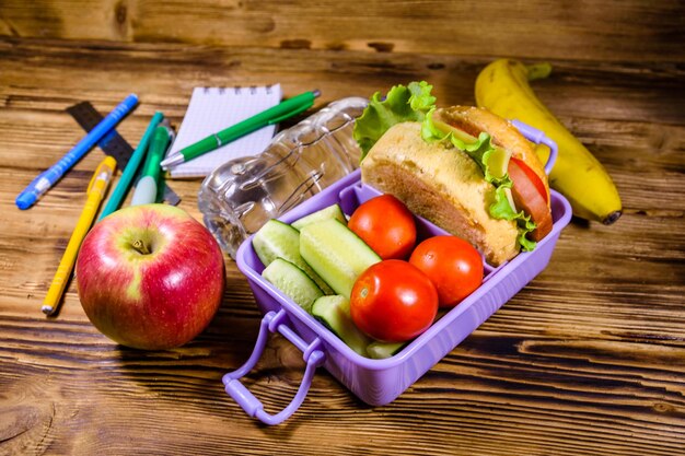 Bottle of water banana ripe apple different stationeries and lunch box with hamburger cucumbers and tomatoes on wooden table