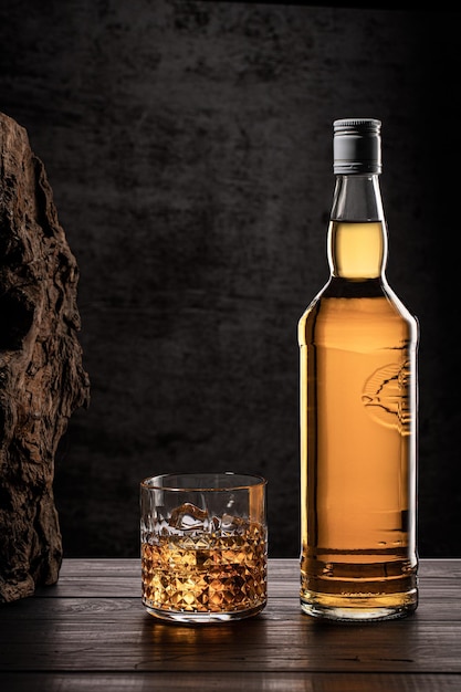 Photo bottle of whisky and glass with ice on wooden table with dark background with copy space vertical format