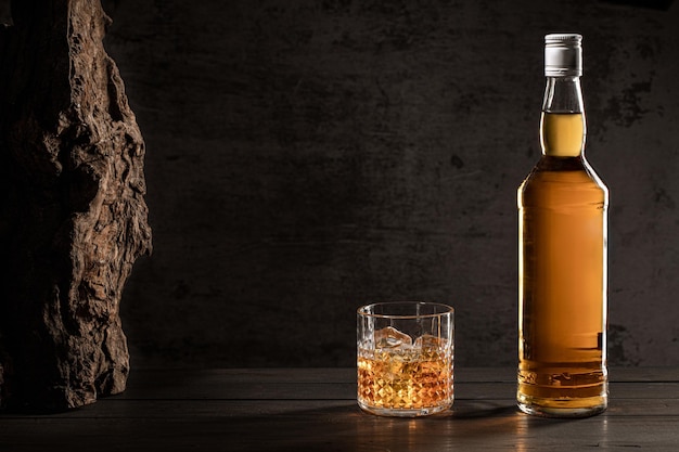 Photo bottle of whisky and glass with ice on wooden table with dark background with copy space