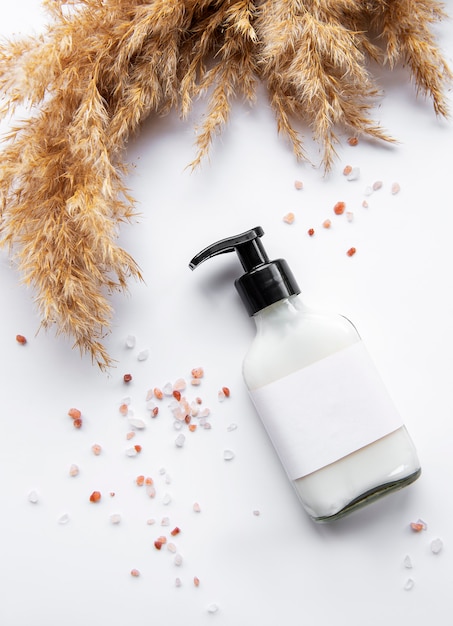 Bottiglia con tappo erogatore ed etichetta pulita, circondata da erba di pampa marrone, su sfondo bianco. concetto di packaging per cosmetici naturali, shampoo, creme. industria della bellezza.