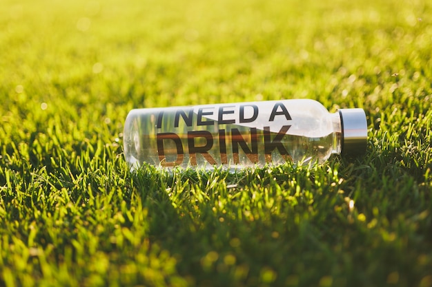 Bottle with fresh clean clear transparent water with text inscription I need drink on green grass on sunshine lawn. Nature texture, green background, wallpaper. Soccer field or sports concept design.