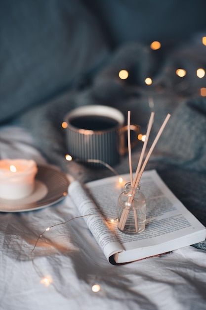 Bottle with liquid home fragrance and wooden bamboo sticks,\
scentes burn candle and cup of black tea over glow lights at\
background close up. winter holiday season. cozy romantic home\
atmosphere.