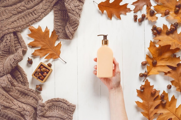 Bottle with liquid soap in female hand on white wooden background with a soft scarf and oak leaves View from above The concept of beauty and care for body and hands