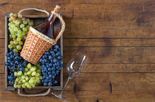 Bottle with red wine, the bunches of ripe grapes in wooden box and a glass on wooden table. Top view, copy-space, rustic style.