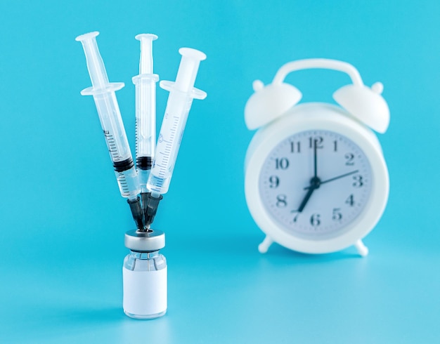 A bottle with a vaccine three medical needles with injections stuck in a bottle an alarm clock on a blue background vaccination against infections