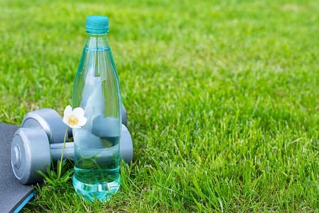 Bottle with water, dumbbells and a mat for sports and yoga on the green grass. Copy space