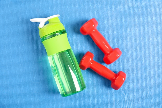 Bottle with water and red dumbbells on blue, top view