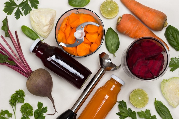 Bottled beet and carrot juices. Root vegetables of beets and carrots. Sliced carrots and beets in blender jar. Cutter. Cabbage and greens. White background. Flat lay