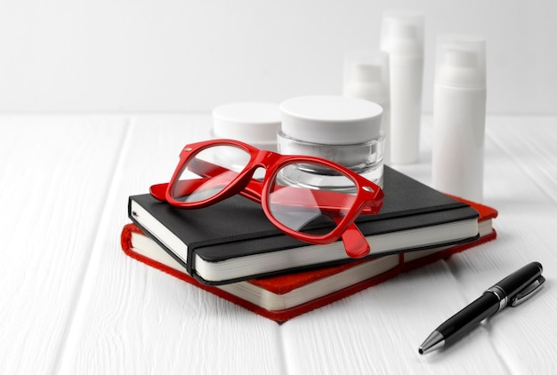 Photo bottles and jars of cream with black and red notepads and glasses on white wooden table