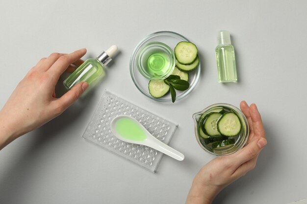 Bottles with cosmetics sliced cucumber female hands and glassware on gray background top view