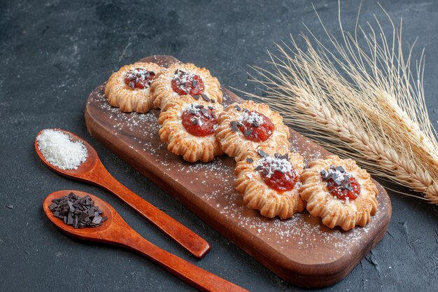 bottom view biscuits with jam on wood board coconut powder and chocolate powder in wooden spoons wheat spikes on dark background