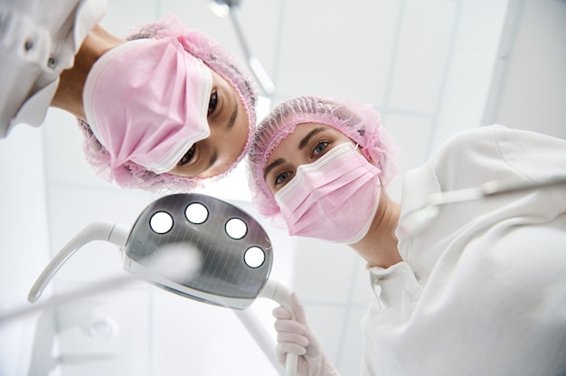 Bottom view of female doctor dentist and assistant adjusting medical light lamp, holding dental instruments and leaning toward a patient for medical examination in white modern clinic. Dental practice