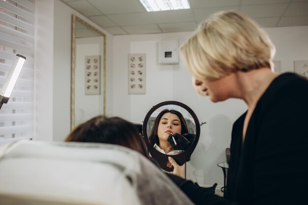 Bottom view of mirror reflection of smiling caucasian girl looking at her beautiful lips while female beautician looking at her after permanent makeup lips procedure in beauty salon Beauty concept