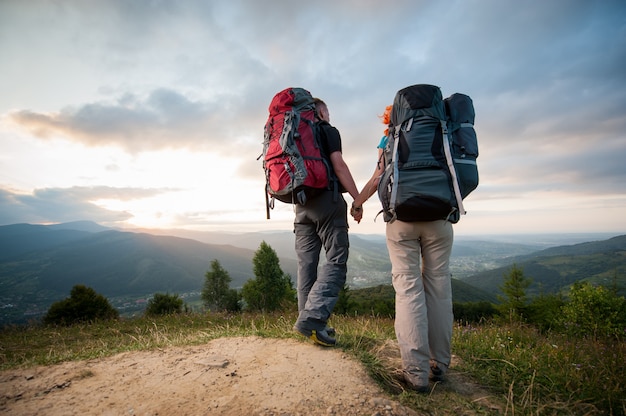 Bottom view pair with backpacks holding hands hiking on the ridge of hill, enjoying the view of beautiful mountains and incredible cloudy sky at sunset