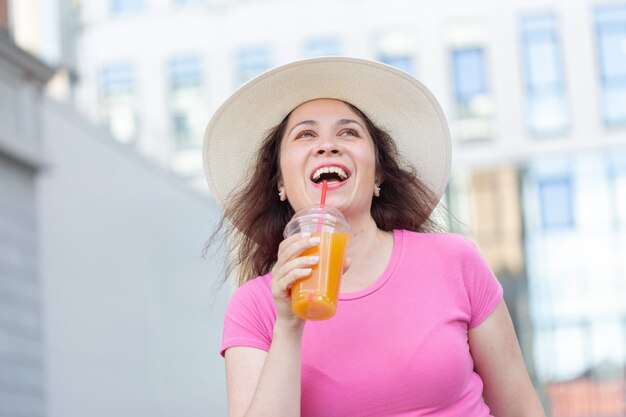 暖かい夏の日にジュースを持って街を歩いている帽子をかぶった若い美しい陽気な女性の底面図の肖像画。週末に街を歩くというコンセプト。