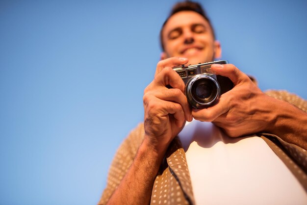 Bottom view of smiling handsome young man with vintage camera take a picture on sky background autumn summer mood background photography