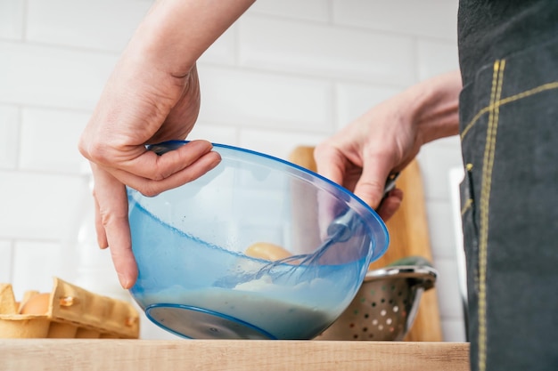 Bottom view of unrecognizable man in black kitchen apron beating eggs in blue plastic bowl with wire whisk on table near steel colander with red yellow bell peppers and open box with eggs Cooking