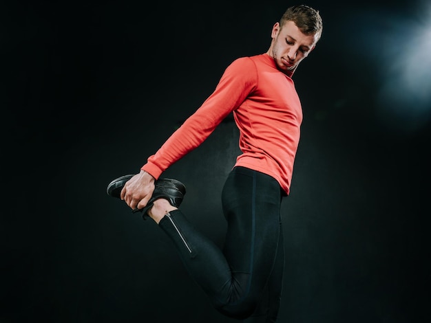 Bottom view of young European man wearing red sportswear and stretching his leg after a heavy workout in dark background Powerful handsome athletic male doing fit Sport motivation Healthy lifestyle