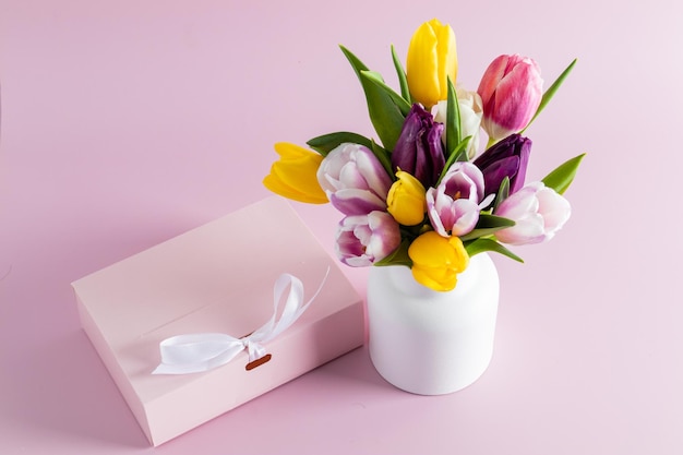 A bouquet of bright spring tulips in a white ceramic vase on a pink background with a gift the concept of a gift and a holiday