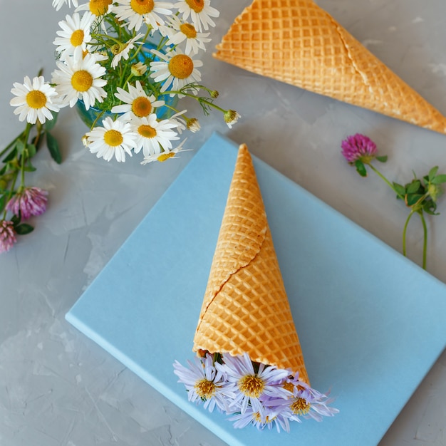 Bouquet of chamomiles in a waffle cone on a gray background and blue book. The flowers are blooming. Wildflowers.
