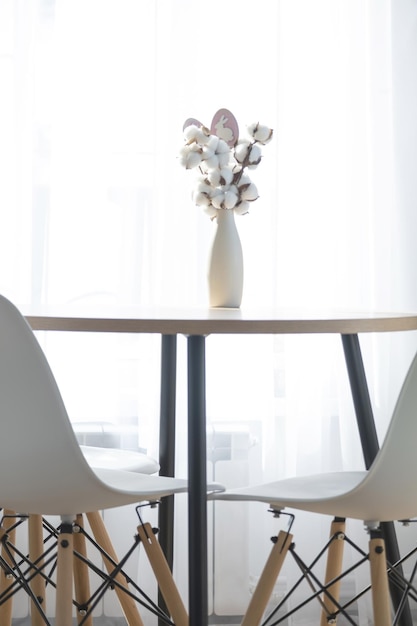 A bouquet of cotton twigs in a vase on a round table with an Easter decor of eggs and an Easter bunny in the light from the window in a minimalist interior