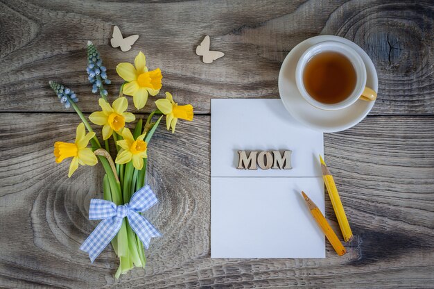 Bouquet of Daffodils and Muscari on wooden table with card, butterflies and cup of tea. Greeting card. Happy Mother's Day. Minimalism, top view, place for text.