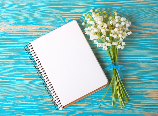 Bouquet of flowers lily of the valley and empty paper notebook on blue rustic table from above, top view, space for text, flat lay.