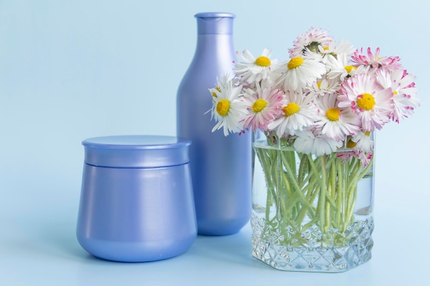 A bouquet of fresh flowers daisies on green stems are in a glass with clean water and two purple jars with cream cosmetics on a blue background