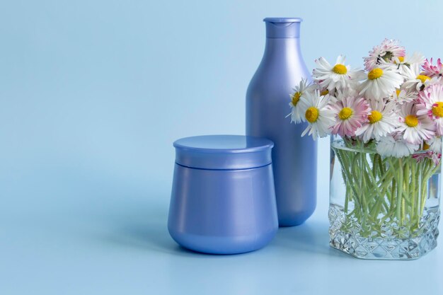 A bouquet of fresh flowers daisies on green stems are in a glass with clean water and two purple jars with cream cosmetics on a blue background