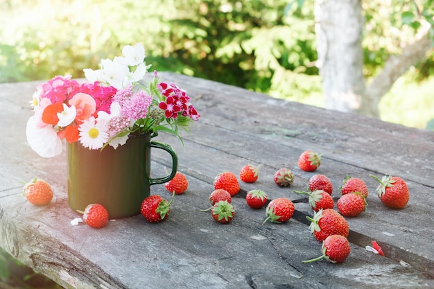 Photo bouquet of garden flowers in green enameled mug and strawberries on old wooden table outdoors