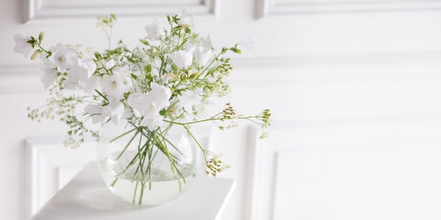 Photo bouquet of gentle bells in vase. morning light in the room. soft home decor, glass vase with white flowers on  white wall background and on wooden table. interior. greeting card. copy space. banner