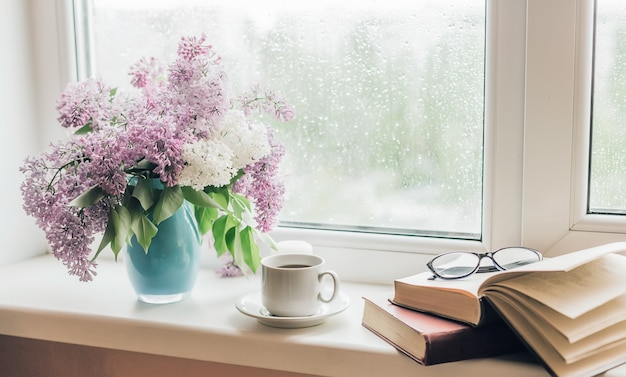 Bouquet of lilacs in a vase,cup of coffee and books on the windowsill.