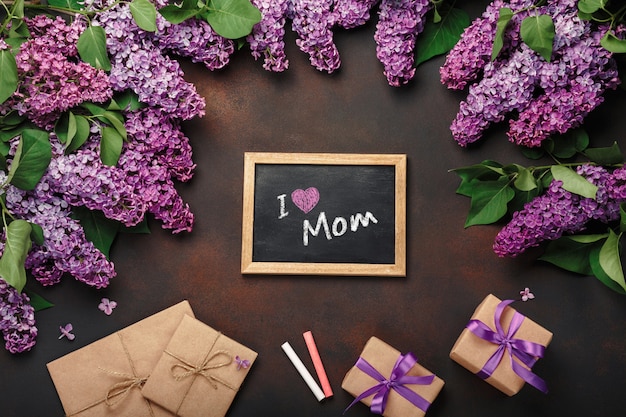 チョークボード、ギフト用の箱、さびた背景にクラフト封筒とライラックの花束。母の日
