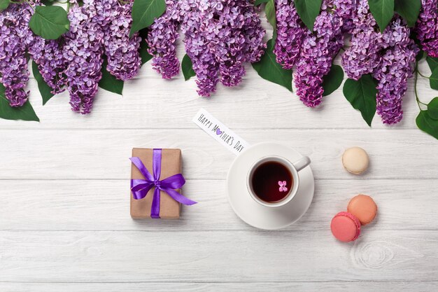 A bouquet of lilacs with cup of tea, gift box, macarons and love note on white boards . Mother's day