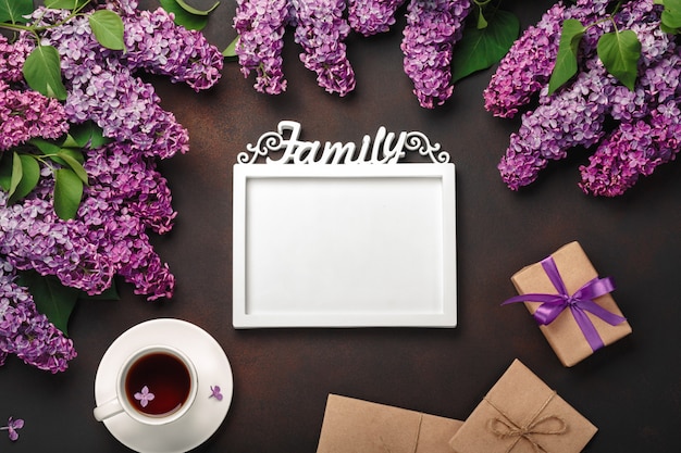 A bouquet of lilacs with cup of tea, a white frame for inscription, gift box, craft envelope, a love note on rusty background