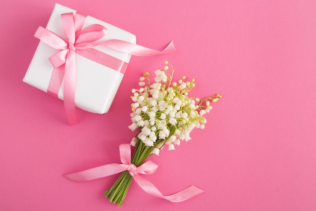 Bouquet of lilies of the valley with pink bow and white gift box on the pink table. Top view. Copy space.