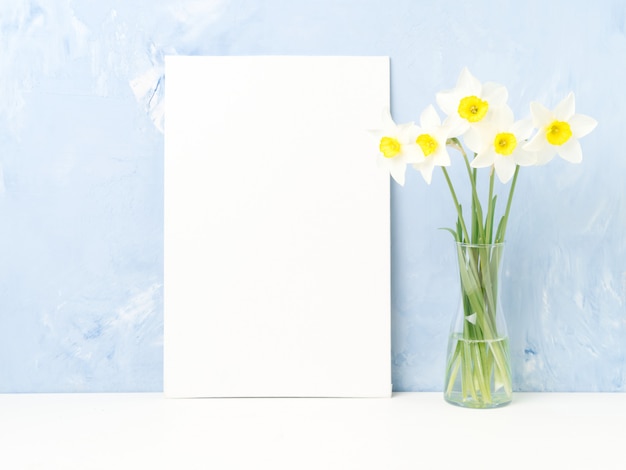 Букет из свежих цветов, чистый лист бумаги, нарциссы со стеклянной вазой на белом столе