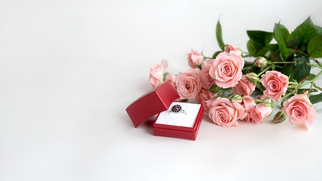 Bouquet of pale pink roses and jeweled engagement ring in opened red jewelry box. Valentine's day celebrating