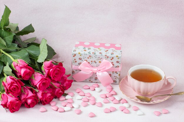 a bouquet of pink roses, a gift in a box, heart-shaped candies and tea in a cup