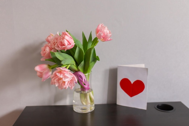 Bouquet of pink tulips in a glass vase with a postcard with a red heart Romantic spring background