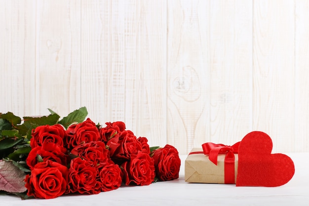A bouquet of red roses, a gift and hearts on a white wooden table 