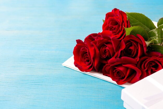 Bouquet of red roses, white gift box, blue wooden background. Copy space. Romantic gift for Valentine's Day holiday