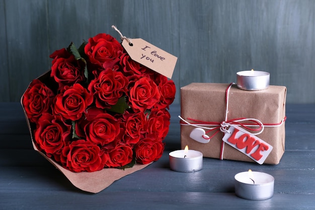 Bouquet of red roses wrapped in paper with present box and candles on wooden background
