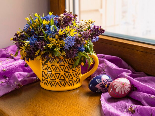 A bouquet of spring flowers with blue snowdrops in a watering can Easter eggs on the windowsill